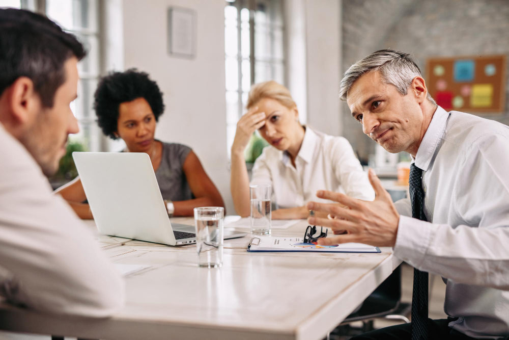 Man gesturing expressively while speaking in a workplace meeting, with three colleagues listening attentively around a conference table.