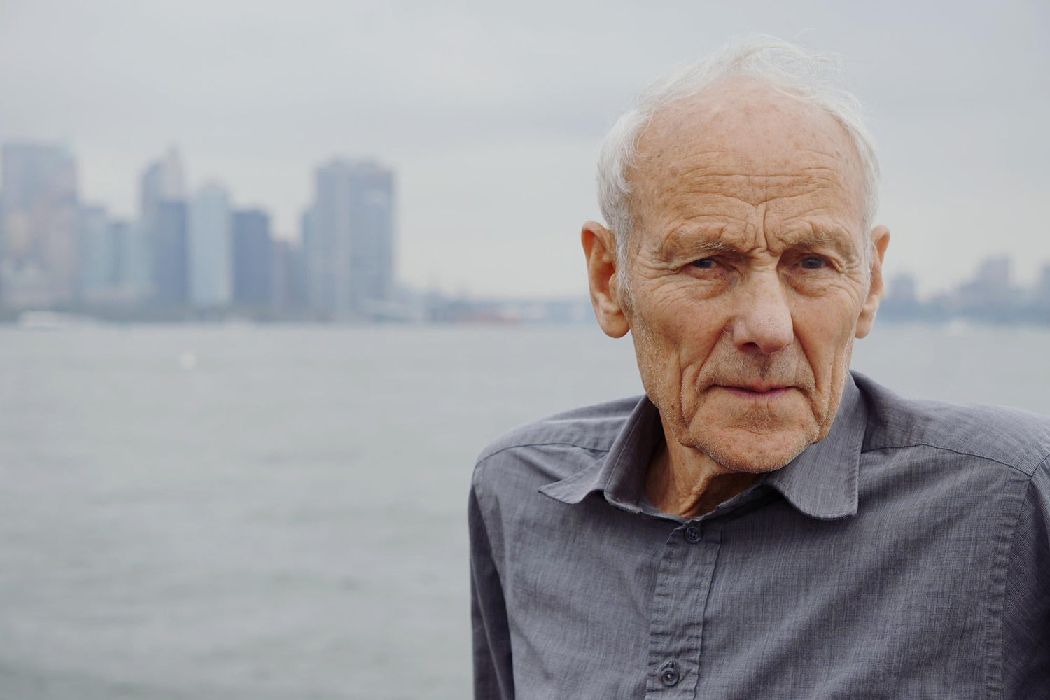 Portrait of a senior Caucasian man standing in a city environment by the sea, neutral expression, grey-hair, wearing a casual shirt, urban skyline and waterfront background.