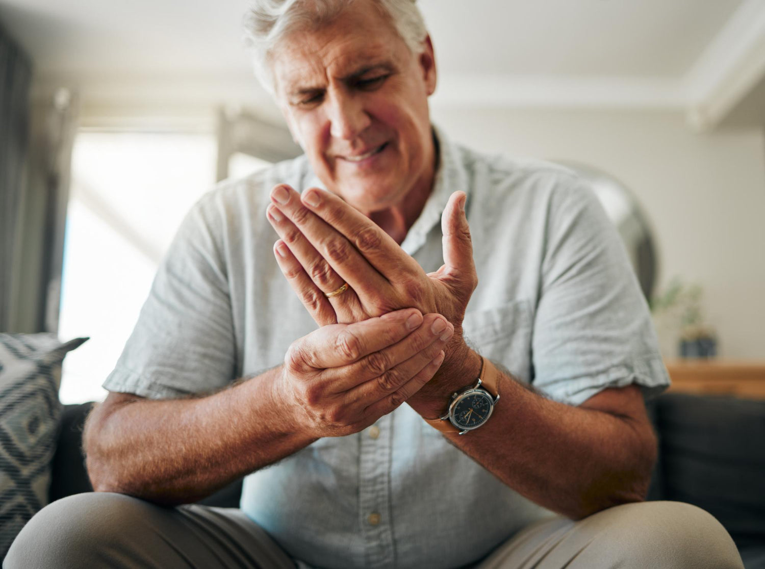Older man sitting on sofa gripping his hand with a pained expression, showing tremors in elderly people.